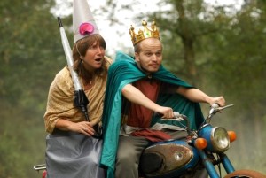 A shocked Queen and King sit atop an old motorbike.