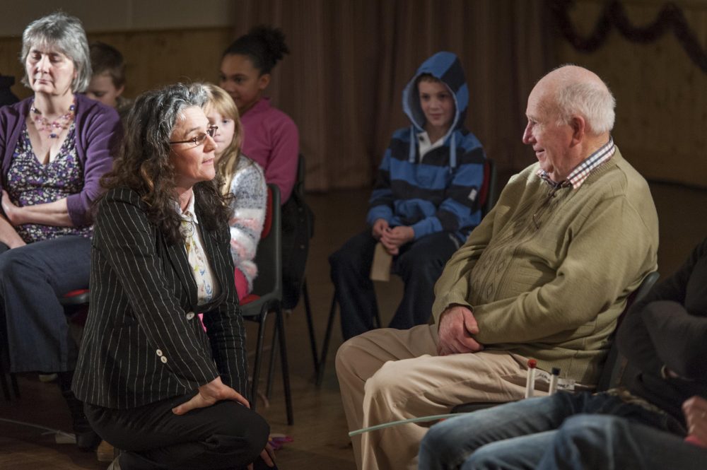 A character in a pine stripe jacker, kneeling and talking to an audience member.