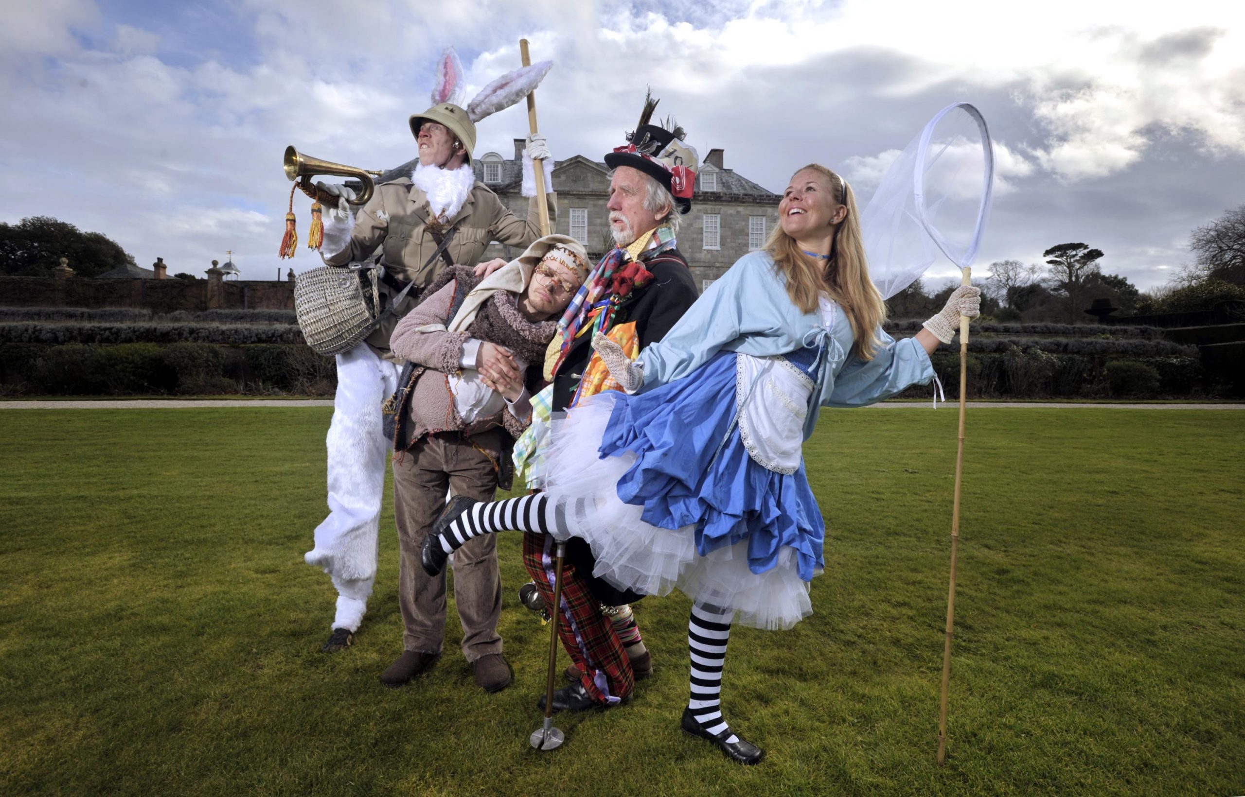 The White Rabbit, Dormouse, Mad Hatter and Alice group together in various poses, outside of a stately home.