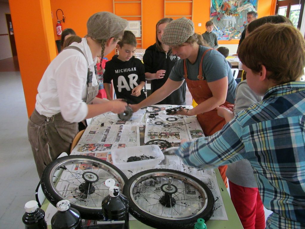 Two flat capped, dungaree wearing characters lead some children in a workshop, painting using bicycle parts.