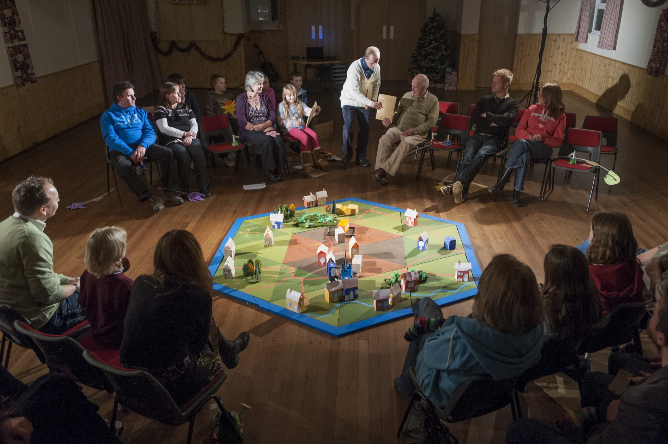 Audience sat in a hall, surrounding a map made of green and brown triangles, with blue edging, place buildings to recreate their village in model form.