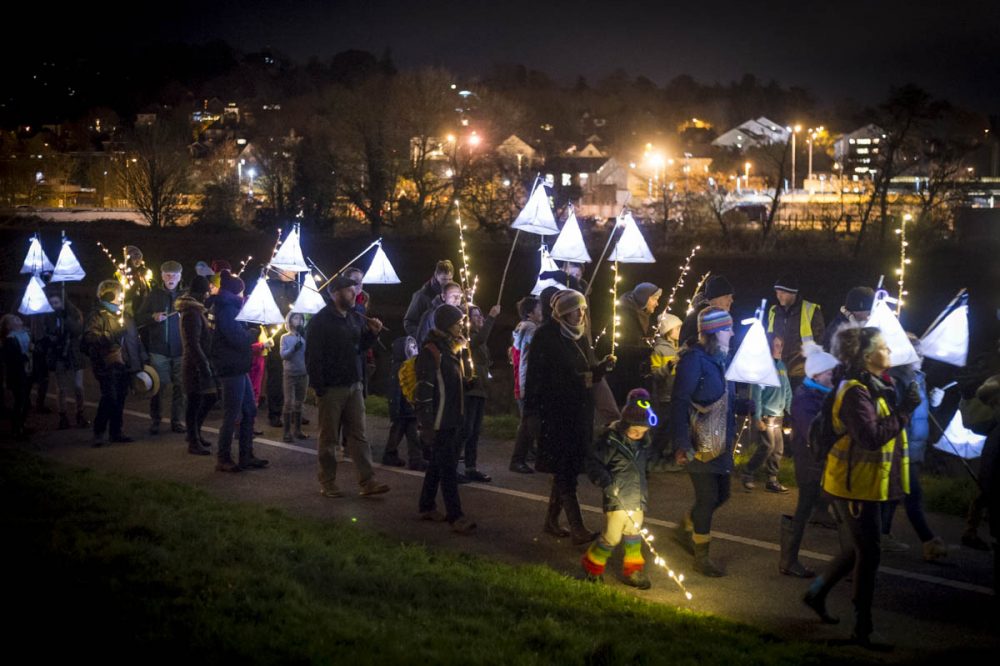 An audience processes alone the canal side at night, holding up handmade lanterns.