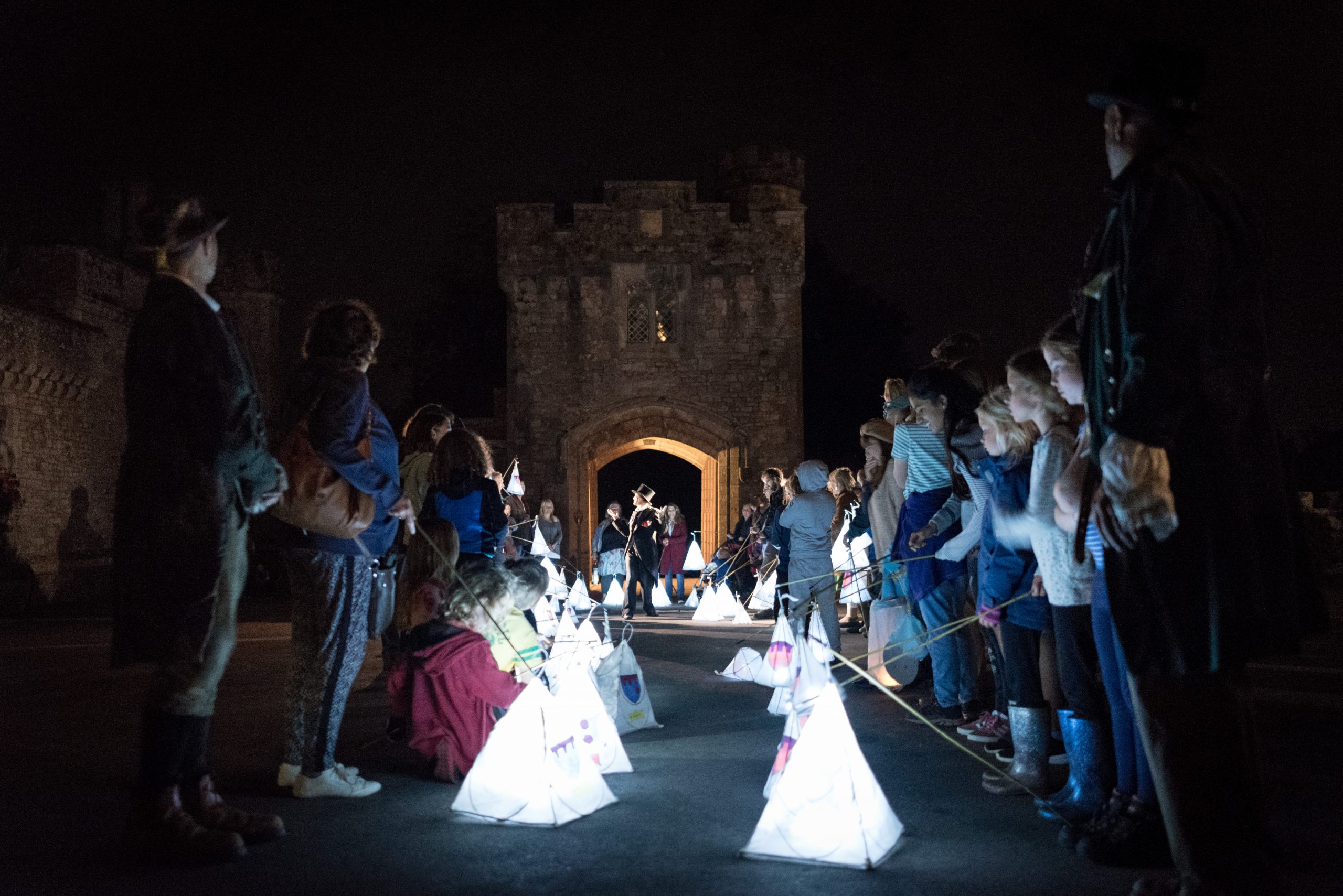 Two lines of audience, with handmade lanterns at their feet, watch on as The Constant Consoler tells them a tale.