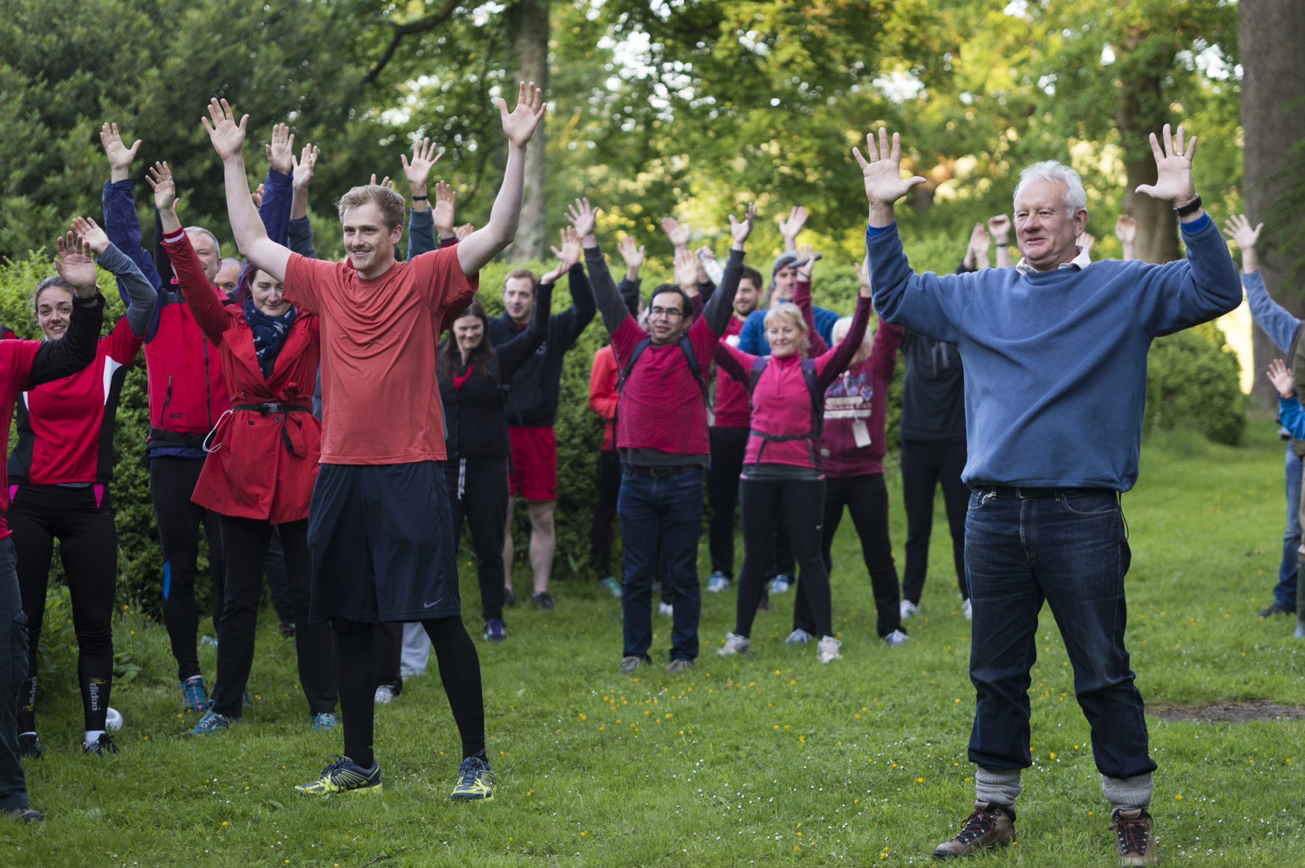 Audience members dressed in red running gear, apart from one chap in a blue jumper and jeans, mid stretch during a warm up routine.