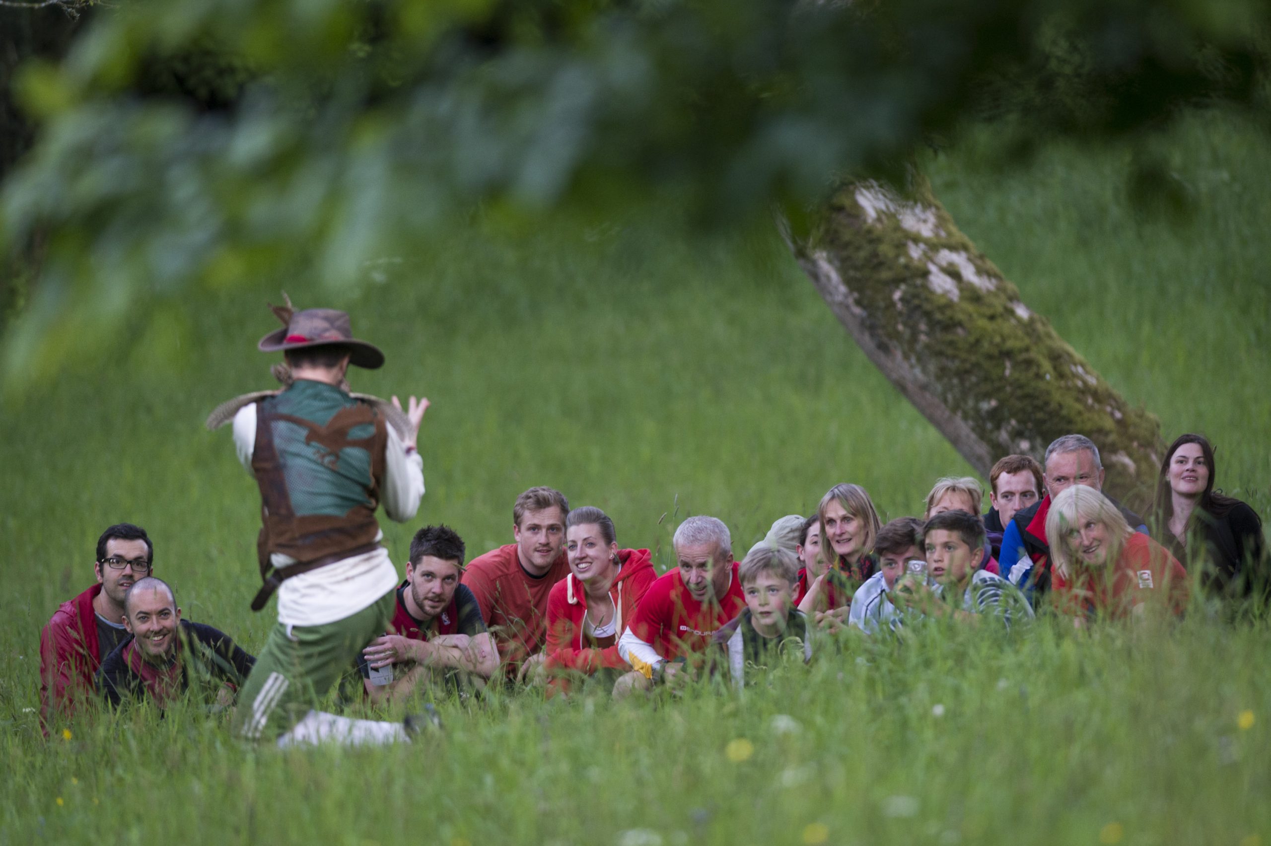 The Huntsman, back to camera, beckons forward the audience who are partially hidden within long grass.