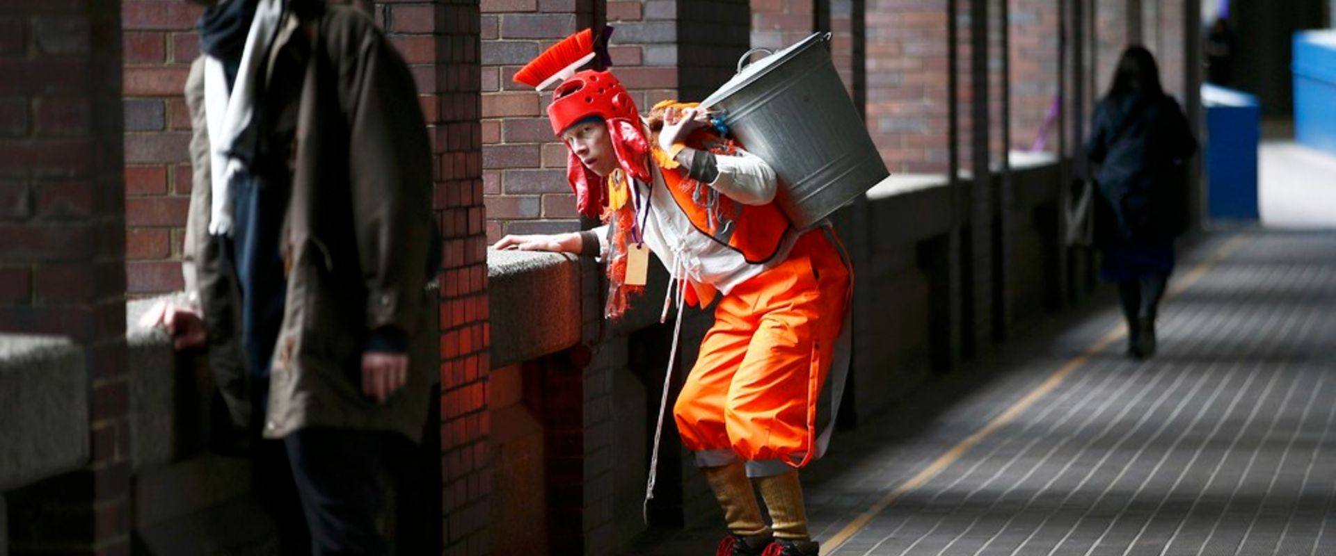 Caliban the Binman, dressed in bright orange hi-vis, and boxing headgear with a broom head atop it, carrying his bin on his back, tries, and fails to look inconspicuous.