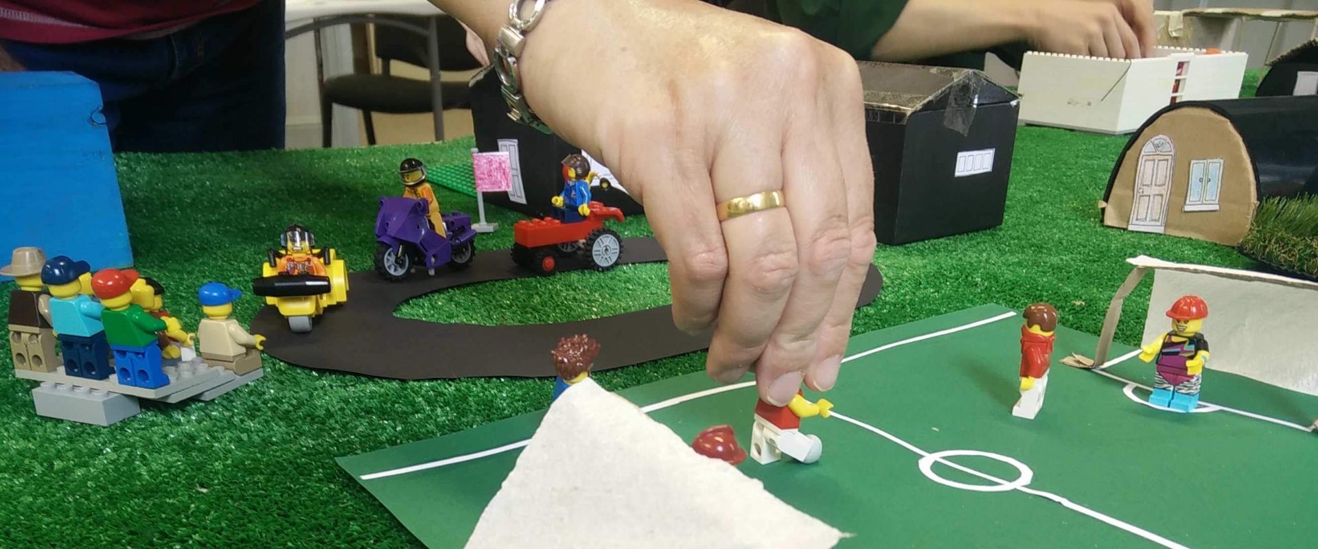 A hand readjusts a Lego figure on a homemade football pitch, the goalkeepers wearing hard hats. Lego speedway happens in the background.