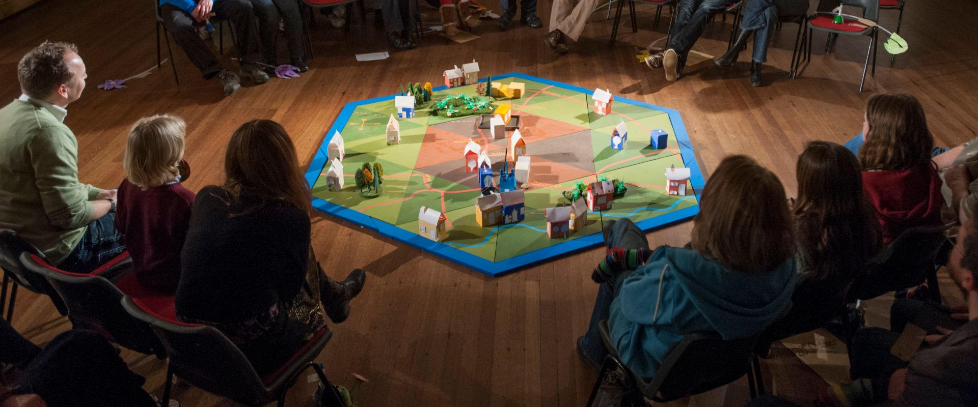 Audience sat in a hall, surrounding a map made of green and brown triangles, with blue edging, place buildings to recreate their village in model form.