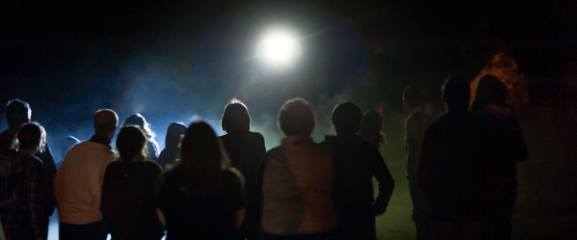 Silhouette of audience at night, watching a glowing light in the sky above them.