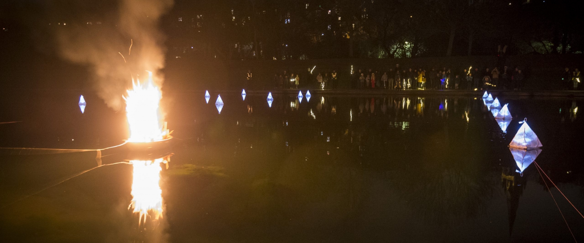 A cardboard ship aflame on the water as fireworks explode.