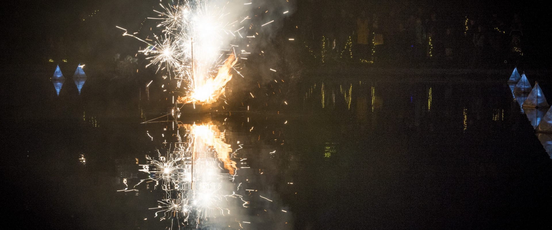 Fireworks spark from a small boat at night, reflected by the river beneath it.