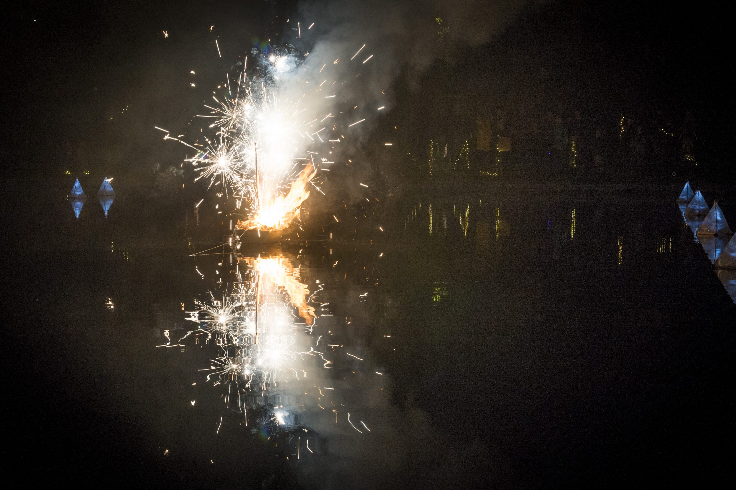 Fireworks spark from a small boat at night, reflected by the river beneath it.