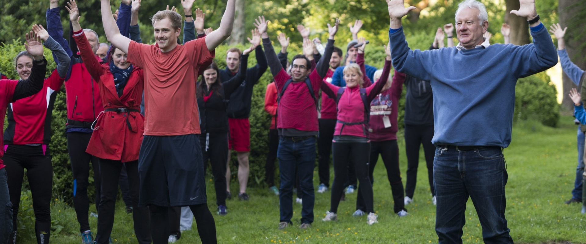 Audience members dressed in red running gear, apart from one chap in a blue jumper and jeans, mid stretch during a warm up routine.
