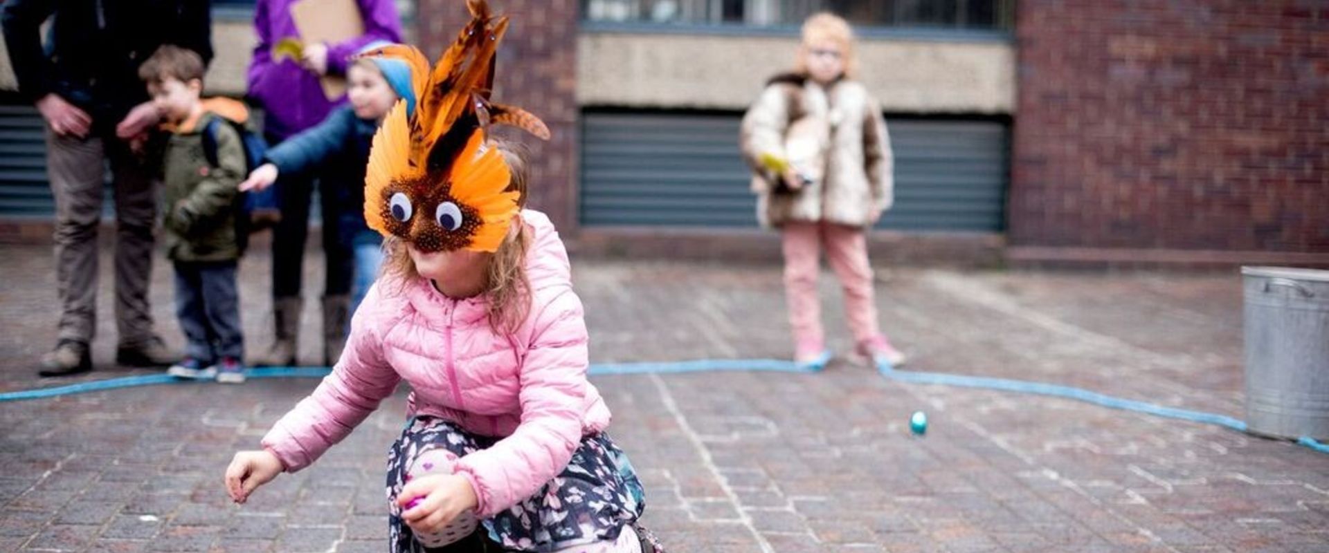 An audience member, wearing a feathered mask with big eyes, searches for something on the ground.